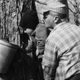 A farmer and his granddaughter inspect a bucket collecting sap from a maple tree in the Catskill Mountains, 1963.